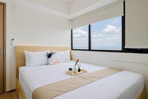 A hotel room with a bed, a window with a city view, a bedside table with a vase and a plant, and a door with a light switch. - Practical 2Br For 3 Pax At Meikarta Apartment (Cikarang)