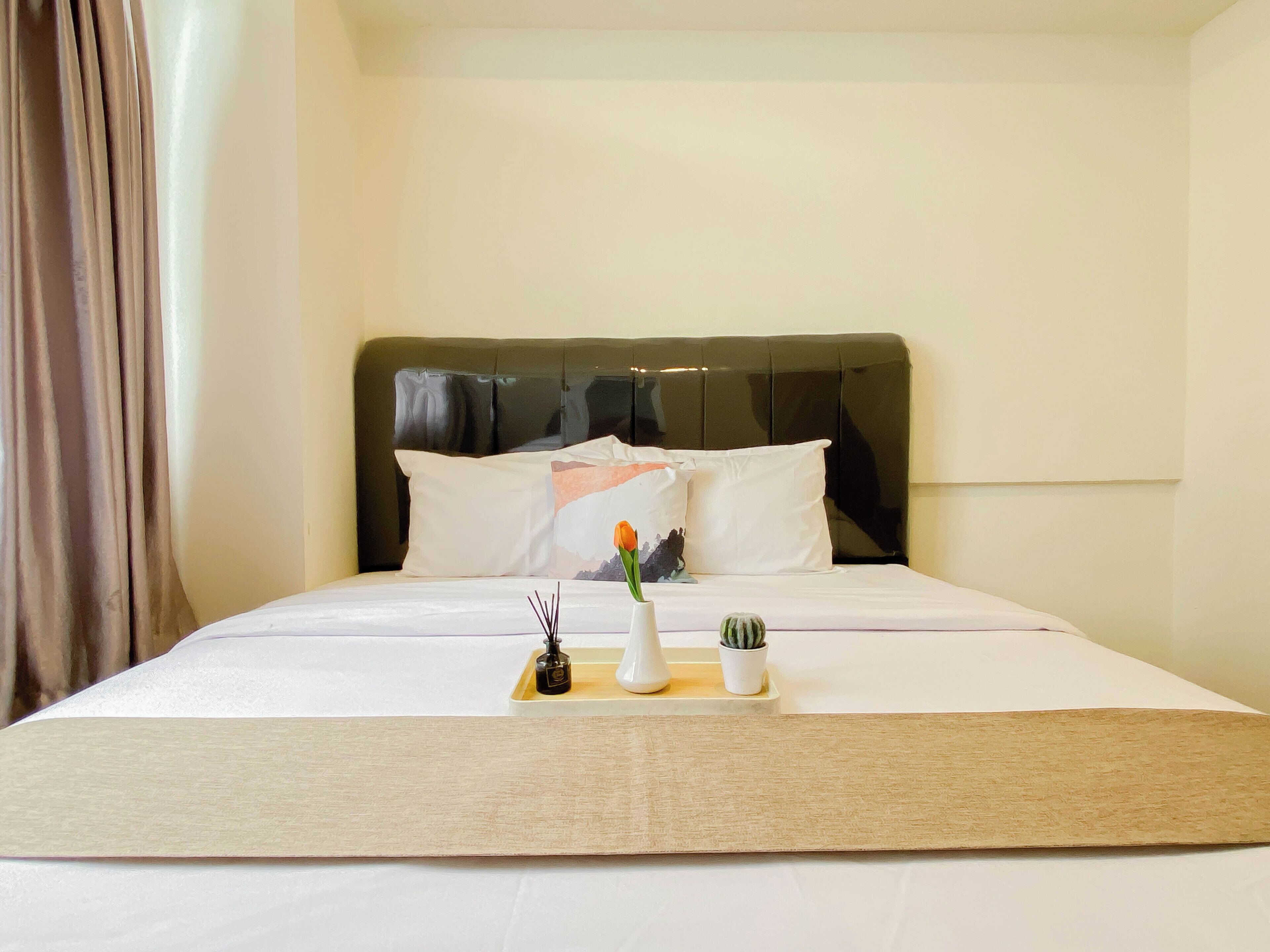 A hotel room with a bed, a headboard, pillows, a bedside table with a vase, and a small plant.