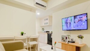 A modern living room with a flat-screen TV, white furniture, a small kitchen area, and a potted plant. - Serene Beige 2Br Space At Apartment Meikarta District 1 (Cikarang)