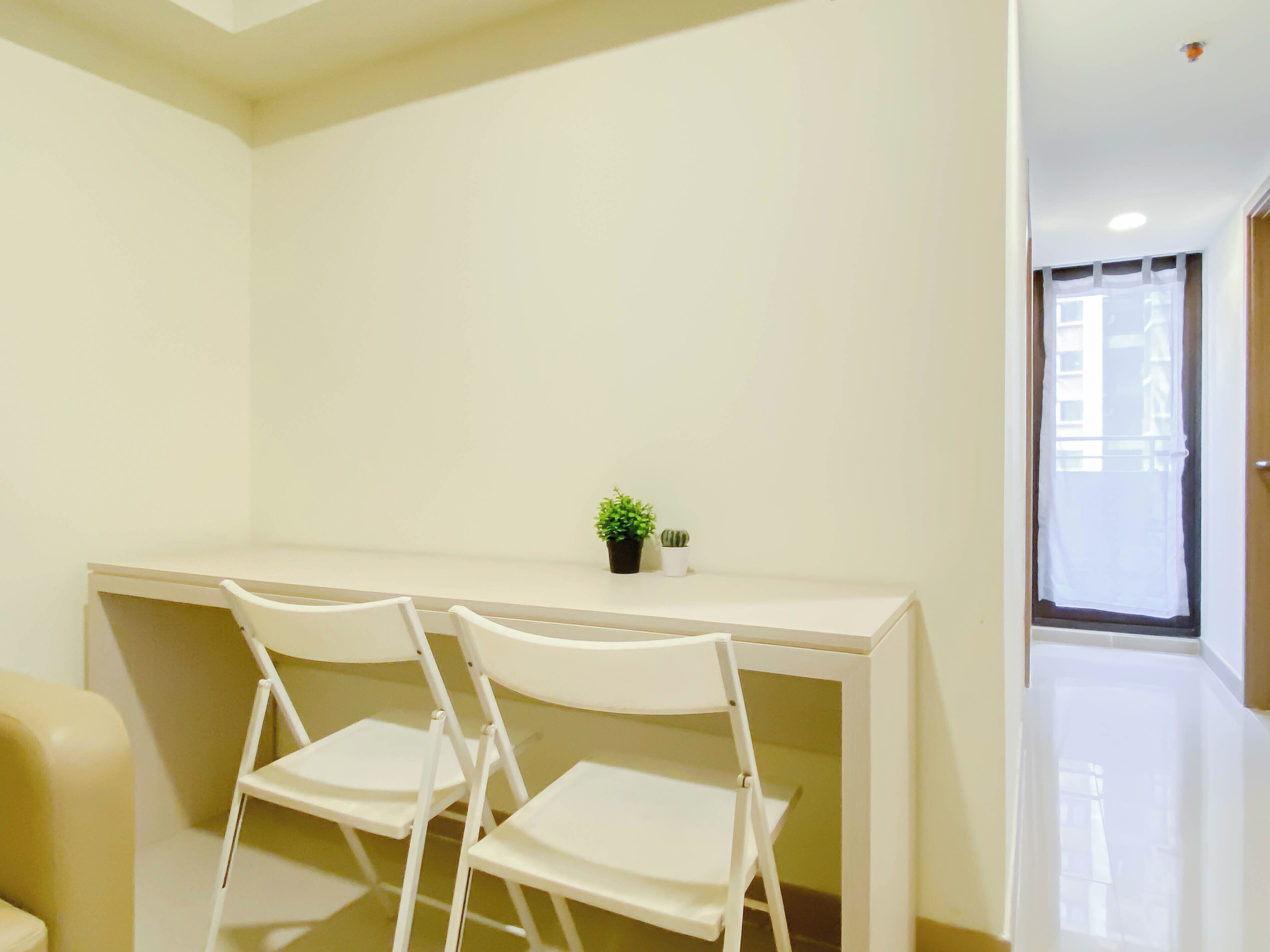 Ein Zimmer mit einem weißen Schreibtisch, zwei weißen Stühlen, einem kleinen Topfpflanzen und einem Fenster mit Vorhang.