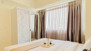 A hotel room with a bed, curtains, a wardrobe, and a window with a view. - Serene Beige 2Br Space At Apartment Meikarta District 1 (Cikarang)