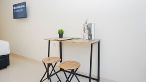 A small, modern room with a bed, a desk with two stools, a potted plant, and a magazine rack. - Comfy And Nice Studio Space At Apartment Meikarta District 1 (Cikarang)