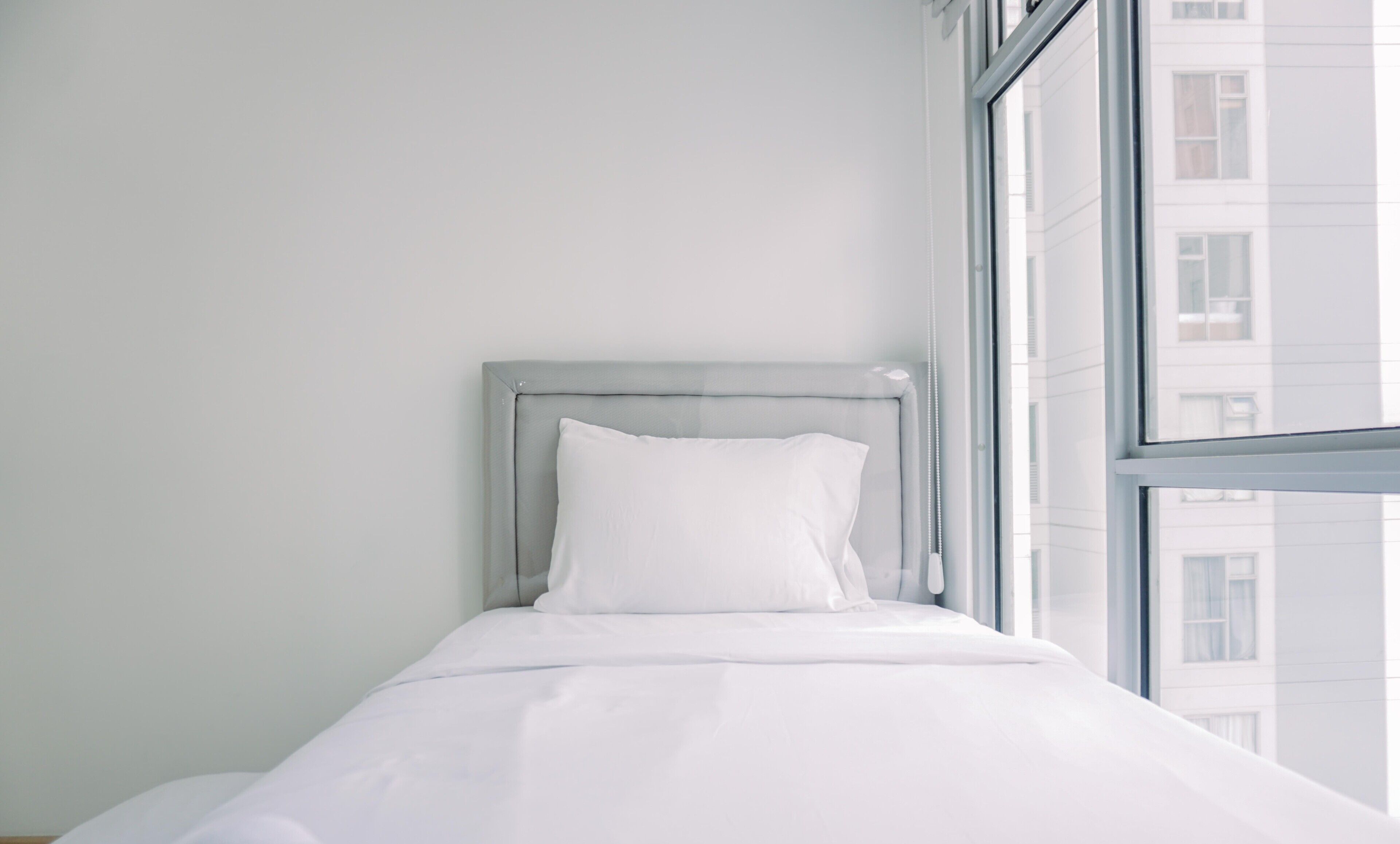 A small, neatly made bed with white bedding in a room with a large window.