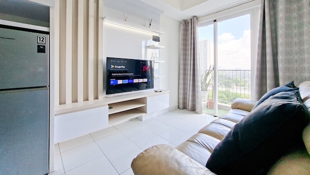 A modern living room with a flat-screen TV, a refrigerator, a sofa, and a balcony with a view. - Comfortable And Homey 1Br Apartment Casa De Parco (Pagedangan)