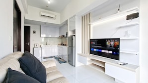 A modern living room with a sofa, a flat-screen TV, a kitchen in the background, and a white tiled floor. - Comfortable And Homey 1Br Apartment Casa De Parco (Pagedangan)