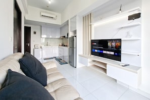 A modern living room with a sofa, a flat-screen TV, a kitchen in the background, and a white tiled floor. - Comfortable And Homey 1Br Apartment Casa De Parco (Pagedangan)