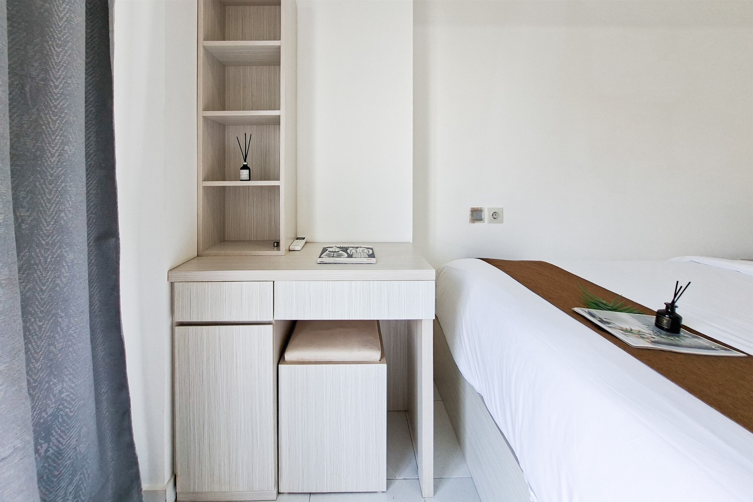 Une chambre d’hôtel avec un lit, un bureau avec des tiroirs, une étagère et une petite table sur laquelle se trouvent un magazine et une bouteille.