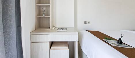 A hotel room with a bed, a desk with drawers, a shelf, and a small table with a magazine and a bottle.