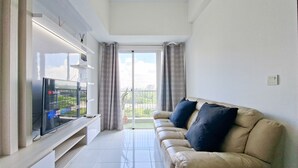 A living room with a flat-screen TV, a beige sofa with dark cushions, a glass coffee table, and a sliding glass door leading to a balcony. - Comfortable And Homey 1Br Apartment Casa De Parco (Pagedangan)