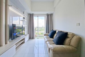 A living room with a flat-screen TV, a beige sofa with dark cushions, a glass coffee table, and a sliding glass door leading to a balcony. - Comfortable And Homey 1Br Apartment Casa De Parco (Pagedangan)