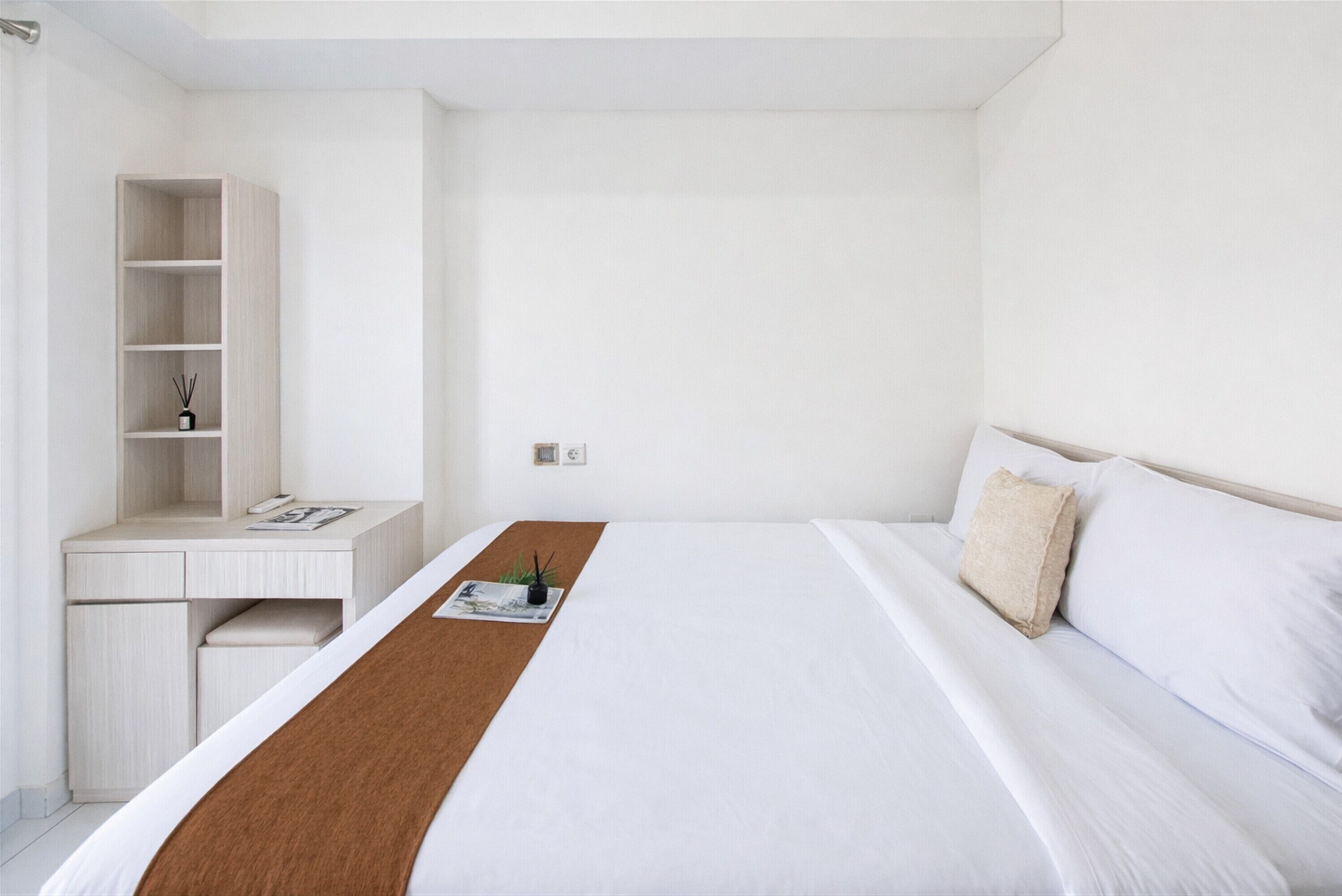 A minimalist hotel room with a bed, a desk, and a shelving unit.