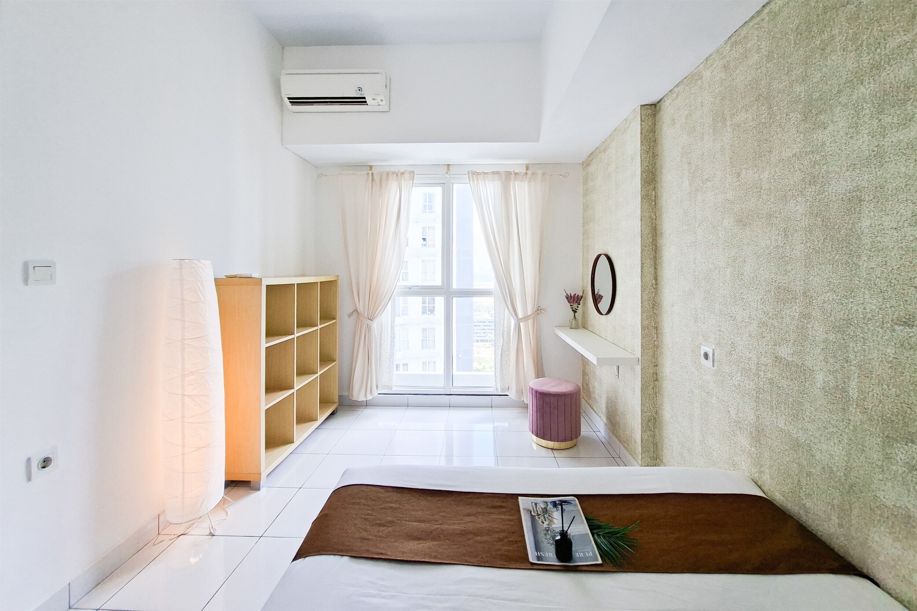 Una habitación de hotel moderna con una cama, un armario de madera, un taburete pequeño, un estante con un espejo y una ventana con cortinas.