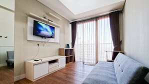 A modern living room with a flat-screen TV mounted on the wall, a grey sofa, wooden flooring, and a large window with curtains. - Calm And Casual Living 1Br At Casa De Parco Apartment (Pagedangan)