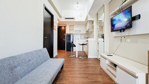 A modern living room with a grey sofa, a flat-screen TV mounted on the wall, and a kitchen area in the background. - Calm And Casual Living 1Br At Casa De Parco Apartment (Pagedangan)
