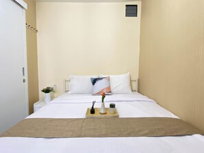Un petit lit bien fait, avec du linge de lit blanc, un plaid beige et un plateau sur lequel se trouvent une bougie, un petit vase et une plante.