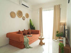 A living room with a sofa, a small side table, a potted plant, and a wall-mounted air conditioner. - Homey 2Br For 3 Pax At Sky House Bsd Apartment (Pagedangan)