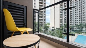 A balcony with a yellow chair and a wooden table, overlooking a swimming pool and high-rise buildings. - Studio Japandi Areum Residence At Sky House Bsd (Pagedangan)
