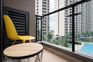 Varanda com cadeira amarela e mesa de madeira, com vista para uma piscina e edifícios altos.