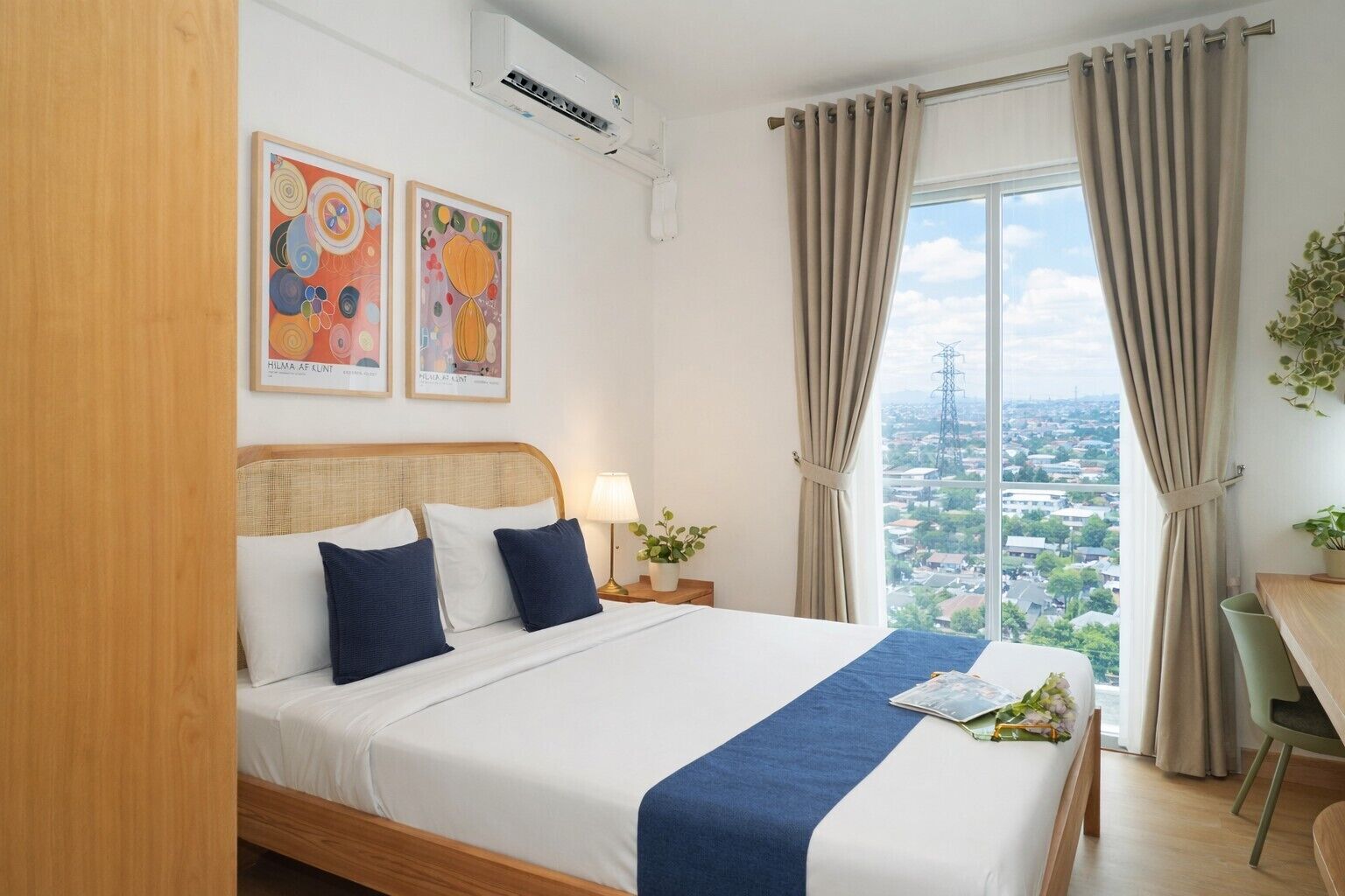 A hotel room with a bed, a nightstand, a lamp, a window with curtains, and two framed artworks on the wall.