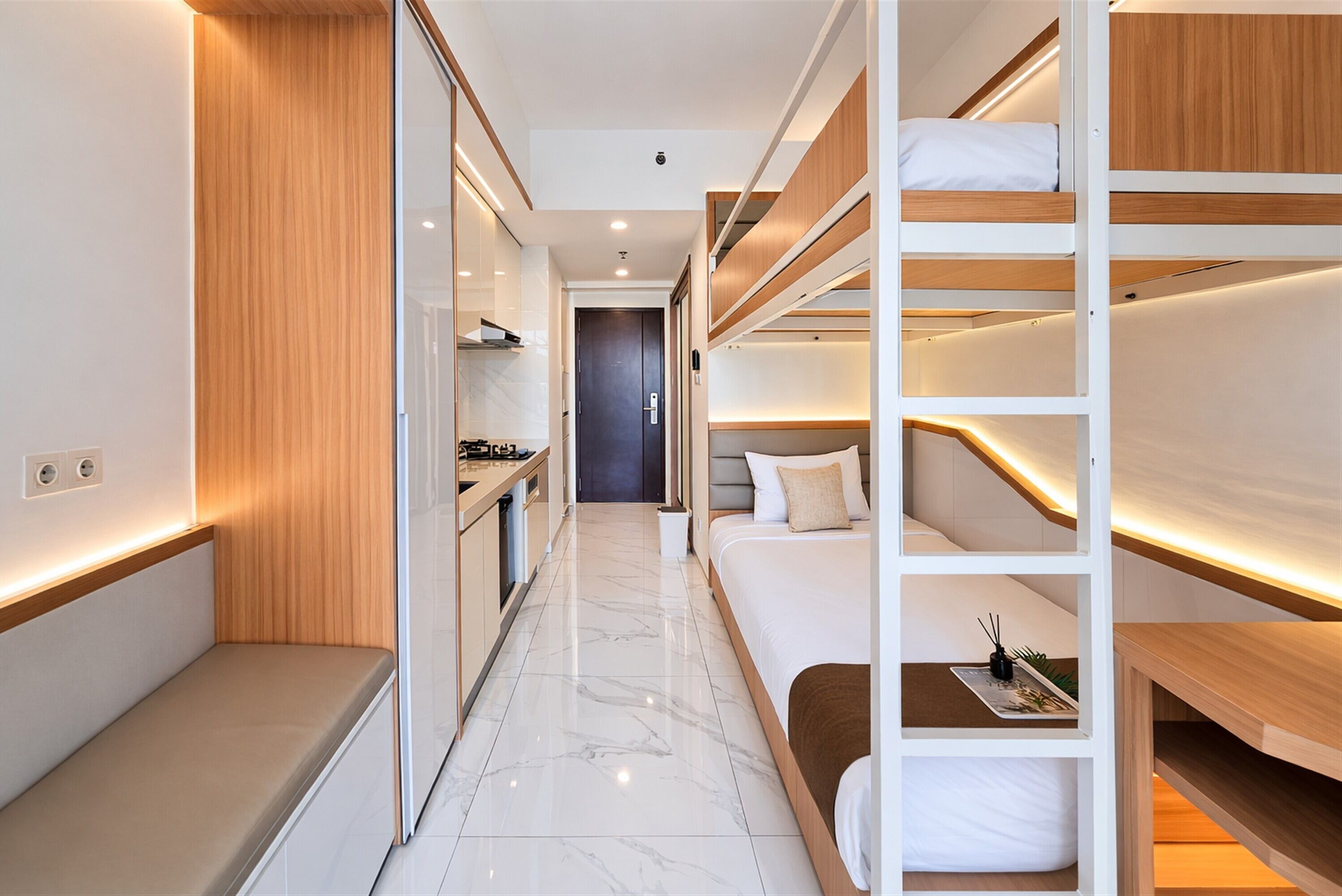 A narrow, well-lit hallway with a bed, a small seating area, and a kitchenette.