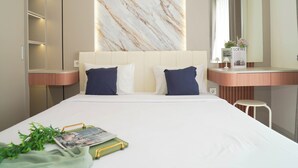 A modern bedroom with a marble wall, a bed with white bedding and blue pillows, a bedside table with a lamp, and a small stool. - Bright And Homey Studio Room At Apartment Sky House Bsd (Balaraja)