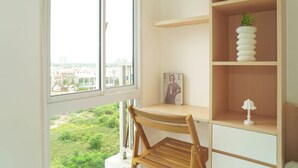 A room with a wooden desk, a chair, a window with a view of buildings, and a shelf with a magazine and a lamp. - Cozy White Studio For 2 Pax At Tokyo Riverside Pik 2 Apartment (Teluknaga)