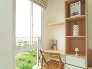A room with a wooden desk, a chair, a window with a view of buildings, and a shelf with a magazine and a lamp. - Cozy White Studio For 2 Pax At Tokyo Riverside Pik 2 Apartment (Teluknaga)