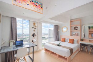 A modern hotel room with a large bed, a desk with a laptop, a view of the city, and a large window.