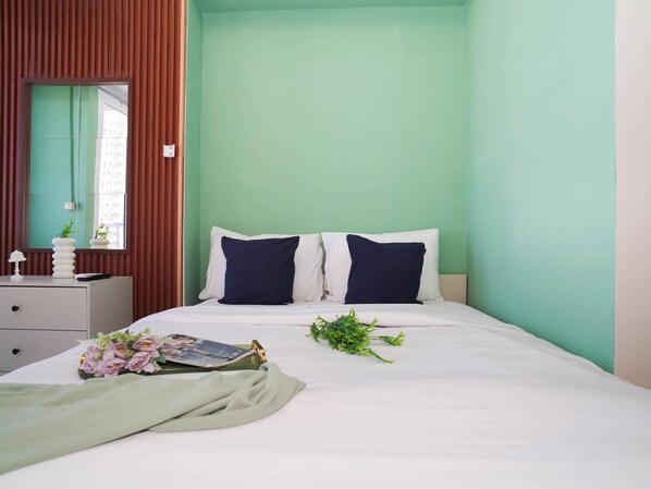 A bedroom with a bed, two pillows, a bedside table with a lamp, and a window with blinds. - Chic Green Studio Room At Apartment Tokyo Riverside Pik 2 (Teluknaga)