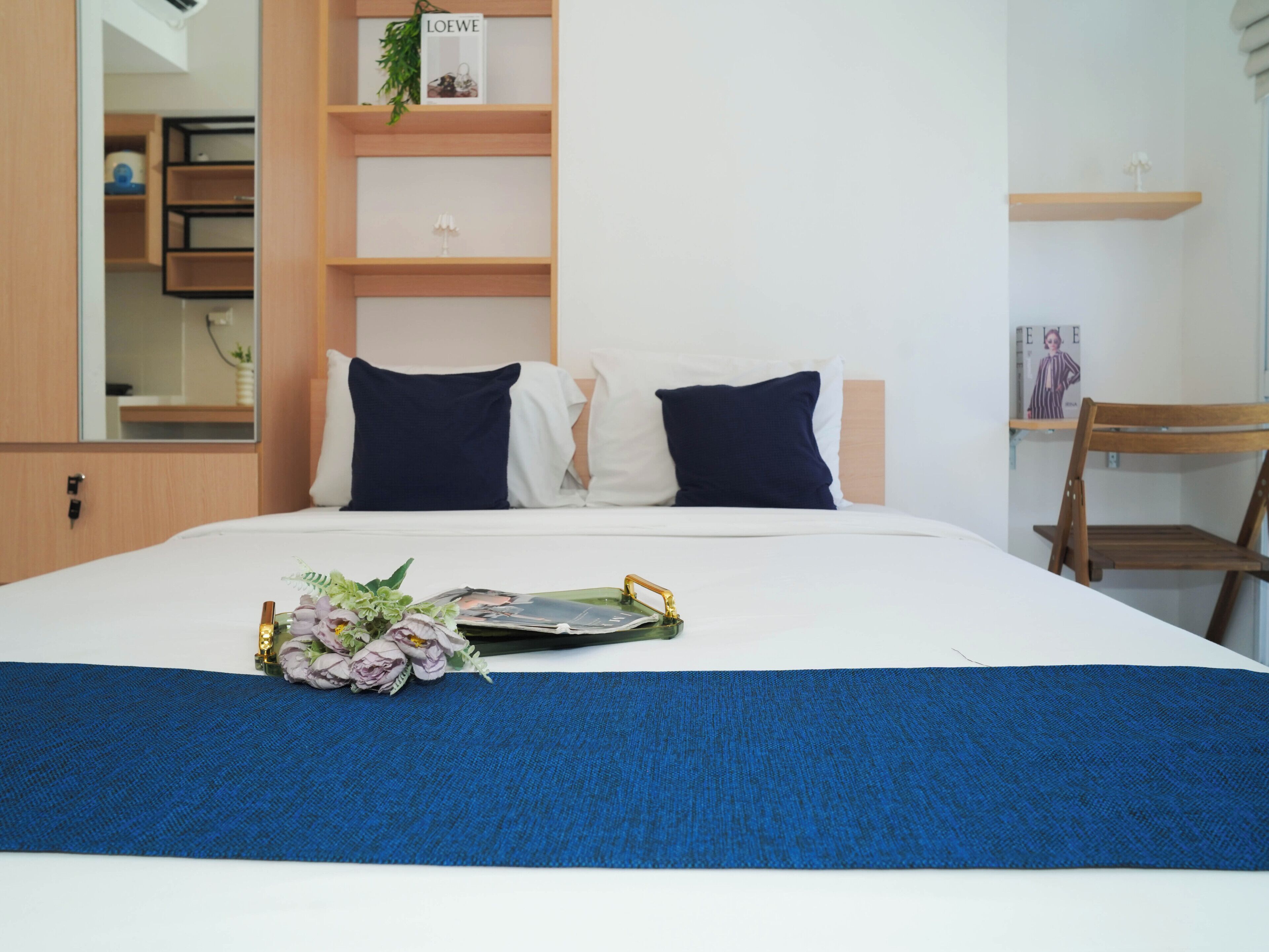 A neatly made bed with a blue bedspread and two pillows, a wooden chair, a small shelf with a plant, and a magazine on the bed.