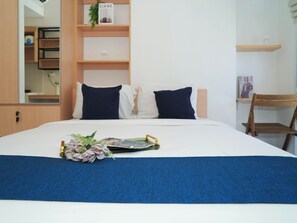 A neatly made bed with a blue bedspread and two pillows, a wooden chair, a small shelf with a plant, and a magazine on the bed. - Best Choice Studio Near Nice At Tokyo Riverside Pik 2 Apartment (Teluknaga)