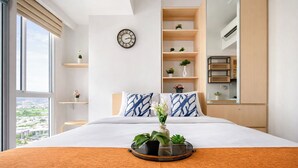 A hotel room with a large bed, a view of the city, a clock on the wall, and shelves with plants. - Practical Studio For 2 Pax At Tokyo Riverside Pik 2 Apartment Near Nice (Teluknaga)