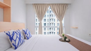Habitación de hotel con una cama grande, dos almohadas azules estampadas, una mesita de noche con un florero y objetos decorativos, y vista a un edificio a través de cortinas translúcidas.