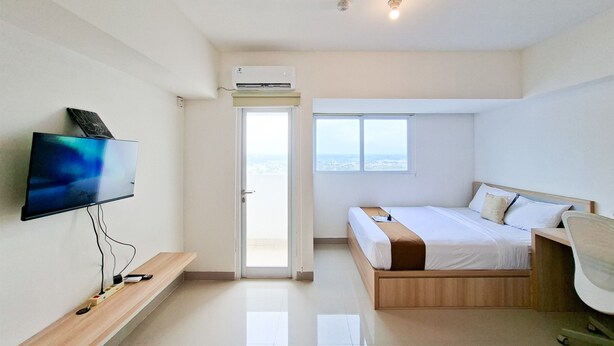 A modern hotel room with a flat-screen TV, a bed, a desk, and a window with a city view. - Comfy And Relaxing Living In Studio At Apartment Cisauk Point (Cisauk)
