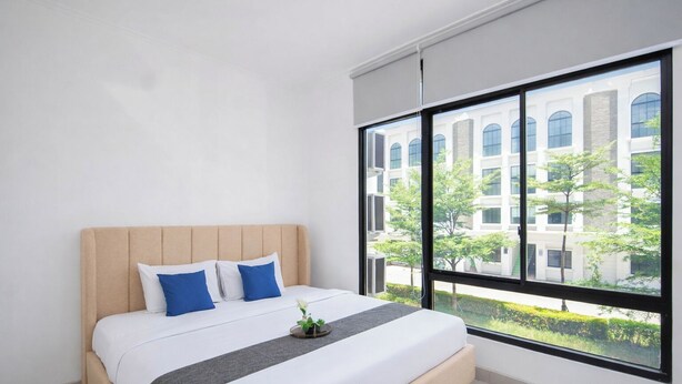 A bedroom with a large window, a bed with white bedding and blue pillows, and a view of a building and greenery outside. - Homey And Modern 2Br At Apartment Green Royal Condo House (Jakarta)