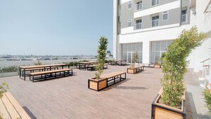 A rooftop terrace with wooden benches and planters, a modern building in the background, and a clear sky. - Japanese Style Studio At Kyo Society Apartment (Surabaya)