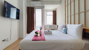 A hotel room with a bed, a TV mounted on the wall, an air conditioning unit, and a window with curtains. - Studio Minimalist And Relaxing At Kyo Society Apartment (Surabaya)