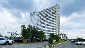 A tall, modern building with a grid of windows, surrounded by parked cars and greenery. - Functional And Cozy Studio At Skyview Medan Apartment (Medan)