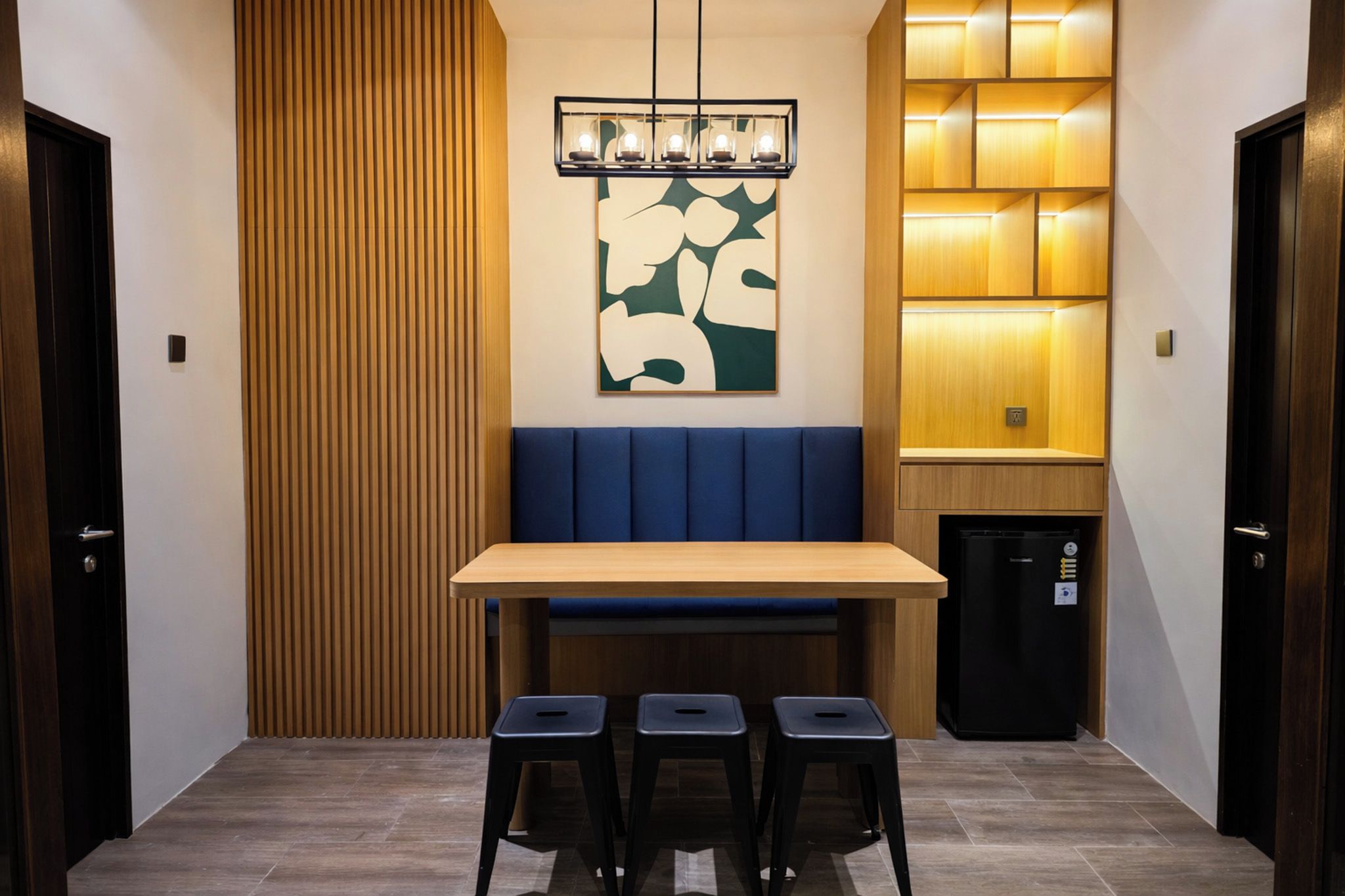 A dining area with a wooden table, blue cushioned bench, three stools, a wall-mounted light fixture, and a refrigerator.
