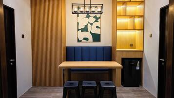 A dining area with a wooden table, blue cushioned bench, three stools, a wall-mounted light fixture, and a refrigerator.