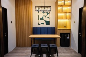 A dining area with a wooden table, blue cushioned bench, three stools, a wall-mounted light fixture, and a refrigerator. - Guru Giri Forest Cabin Villa 2BR with Pool Access (Srimulyo)