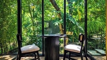A room with a glass wall, two chairs, a round table, and a view of dense greenery.