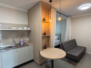 A compact living space with a kitchenette, a round wooden table, a grey sofa, and a wooden cabinet with shelves. - Ravello Residencial – Comfort and tranquility in Foz do Iguaçu (Foz do Iguaçu)
