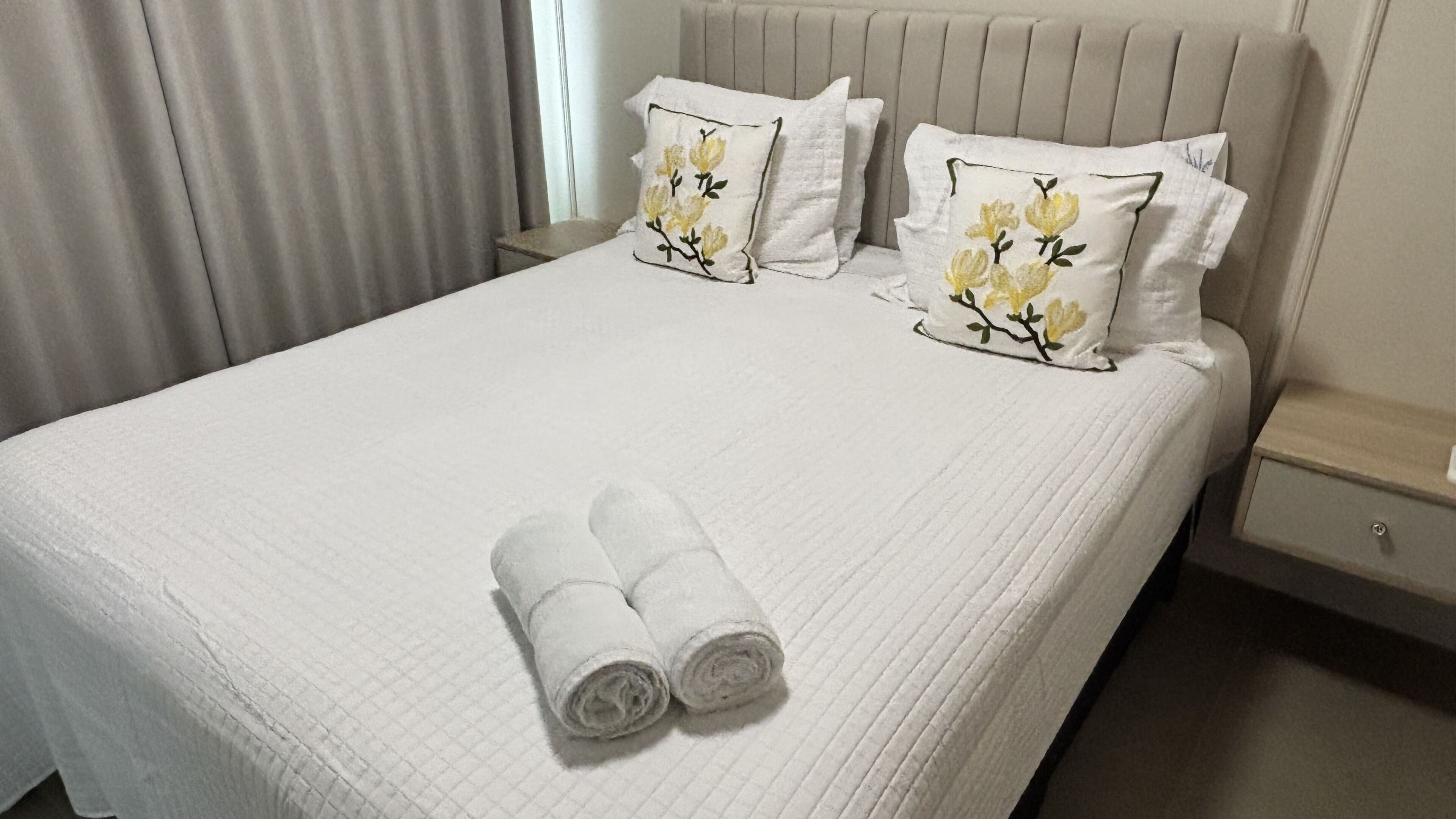 Cama bem arrumada com duas almofadas e dois rolinhos de toalha, uma mesinha de cabeceira e um criado-mudo.
