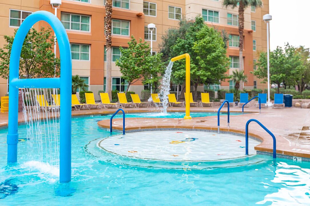 A swimming pool with a water feature, surrounded by yellow chairs and palm trees.