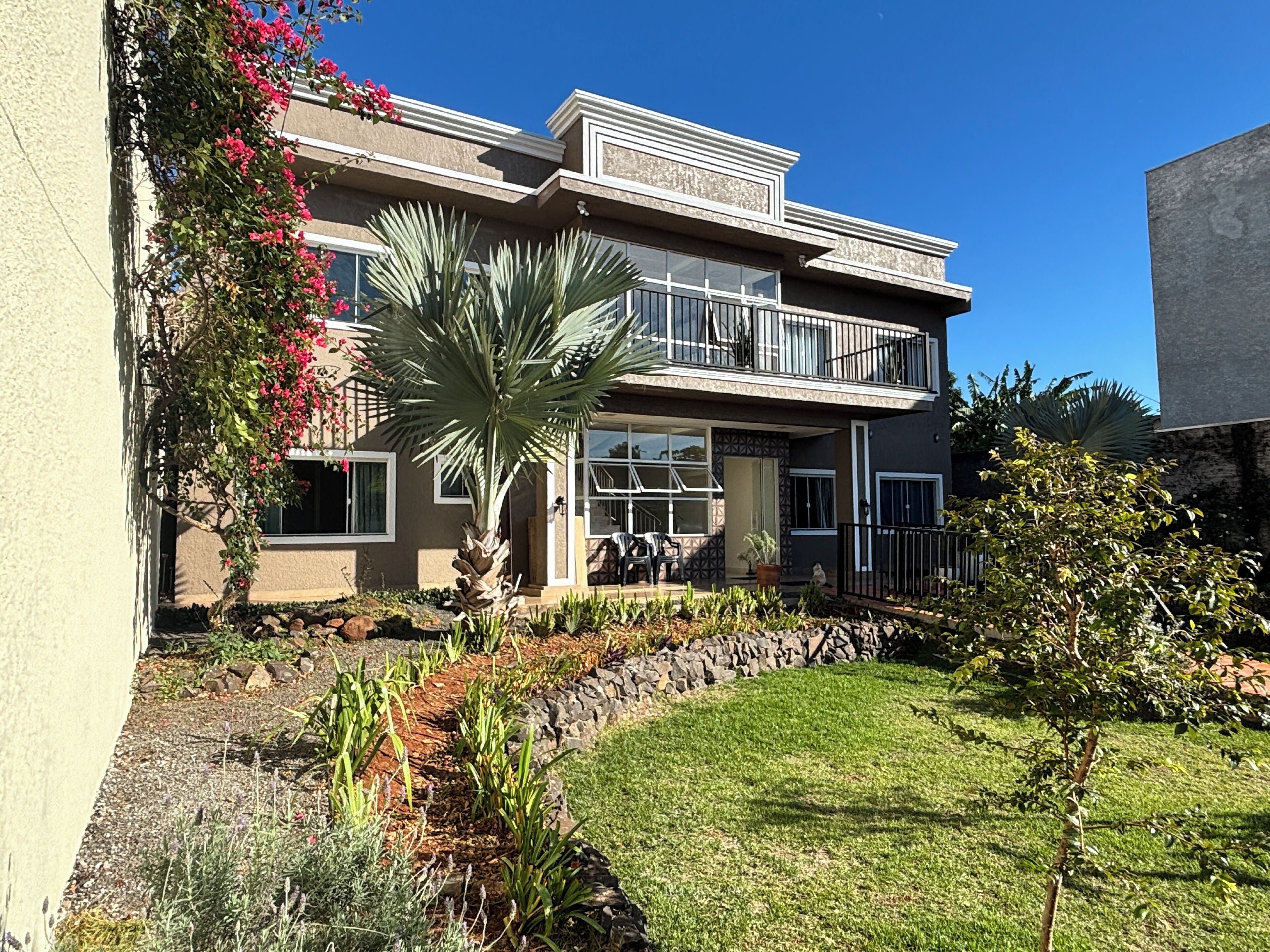 A two-story building with a balcony, a garden with various plants, and a clear blue sky.