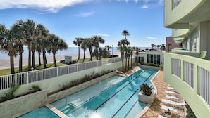 A pool area with multiple swimming lanes, palm trees, and a view of the beach. - Ocean Grove 1003 - Pelican Sky (Galveston)