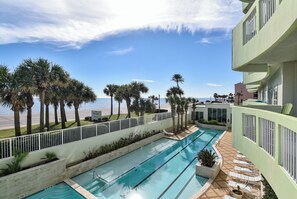 A pool area with multiple swimming lanes, palm trees, and a view of the beach. - Ocean Grove 1003 - Pelican Sky (Galveston)