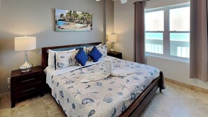 A bedroom with a bed, two bedside tables, a ceiling fan, a painting, and a window with curtains. - Ocean Grove 1003 - Pelican Sky (Galveston)