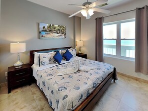 A bedroom with a bed, two bedside tables, a ceiling fan, a painting, and a window with curtains. - Ocean Grove 1003 - Pelican Sky (Galveston)